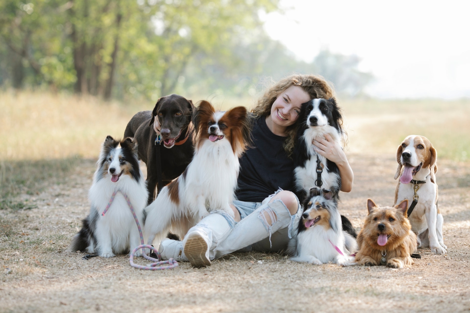 Een jonge vrouw die glimlacht naar de camera en omringt is door verschillende honden